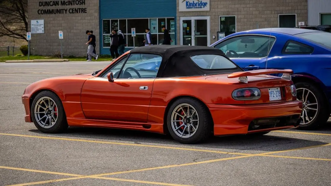 Red Mazda MX-5 with aftermarket body kit parked in urban lot near a community center